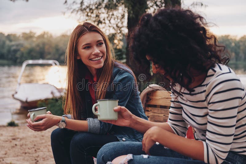 Good talk with friend. Two young beautiful women in casual wear smiling and talking while enjoying camping near the lake. Good talk with friend. Two young beautiful women in casual wear smiling and talking while enjoying camping near the lake