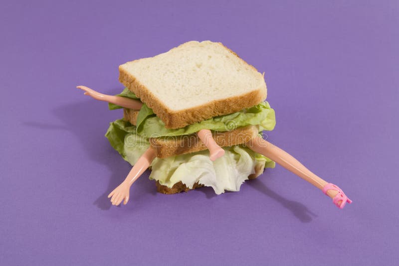 Parts of a doll`s body in a sandwich with salad and soft bread on a minimal background color. pop fun and quirky cannibalism. Parts of a doll`s body in a sandwich with salad and soft bread on a minimal background color. pop fun and quirky cannibalism