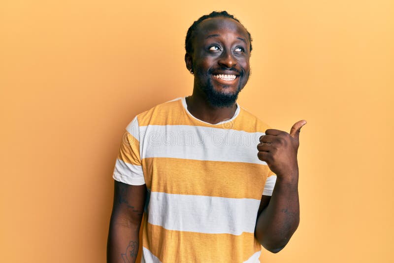 Handsome young black man wearing casual yellow tshirt smiling with happy face looking and pointing to the side with thumb up. Handsome young black man wearing casual yellow tshirt smiling with happy face looking and pointing to the side with thumb up