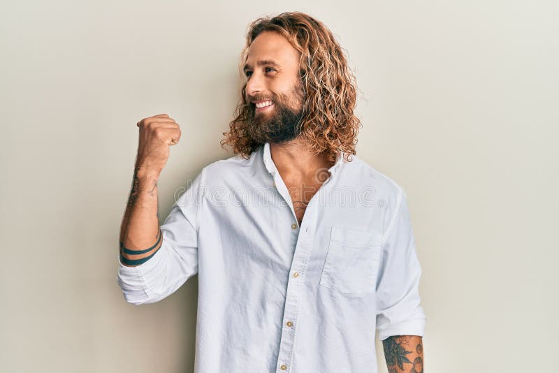 Handsome man with beard and long hair wearing casual white shirt pointing thumb up to the side smiling happy with open mouth. Handsome man with beard and long hair wearing casual white shirt pointing thumb up to the side smiling happy with open mouth