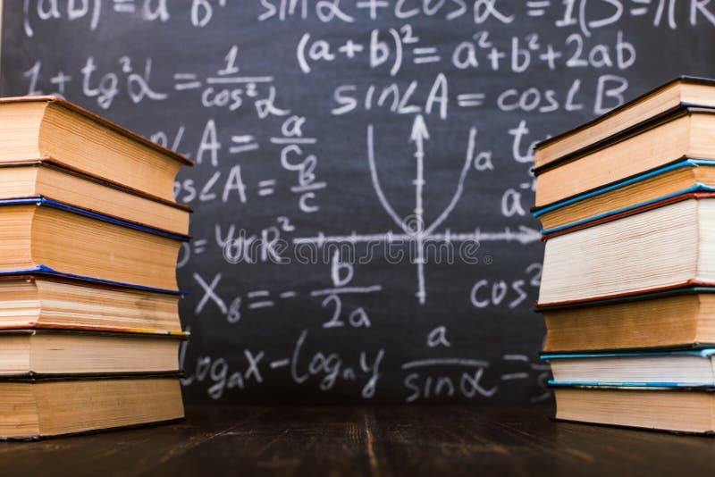 Books on a wooden table, against the background of a chalk board with formulas. Teacher&x27;s day concept and back to school