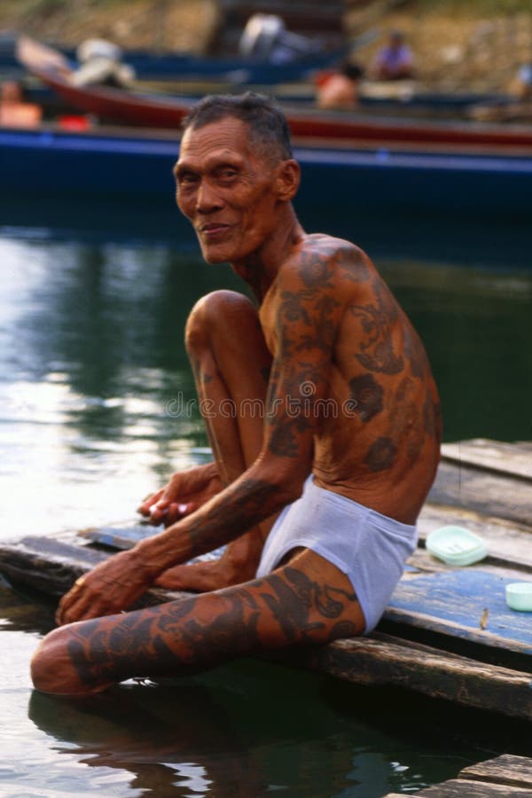 Borneo: Older Iban Headhunter with tatoo`s at the river sitting in the rain forest of Sarawak