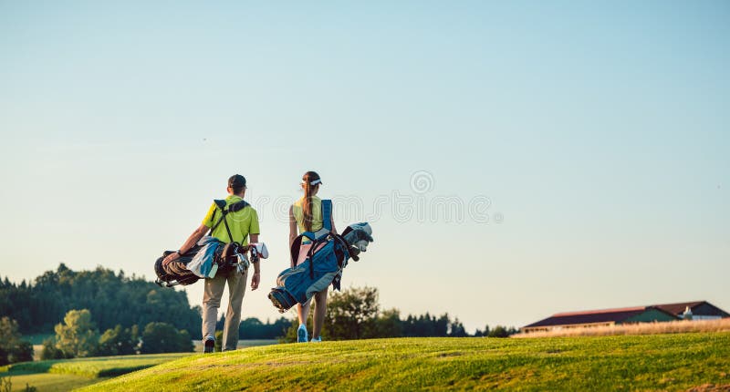 Borse di trasporto del supporto delle coppie felici verso il campo da golf in un giorno soleggiato immagini stock libere da diritti Borse di trasporto del supporto delle coppie felici verso il campo da golf in un giorno soleggiato immagini stock libere da diritti