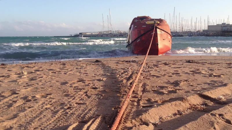 Bote de salvamento anaranjado en la orilla de mar Mediterráneo con las ondas tempestuosas y puerto en el backgroud