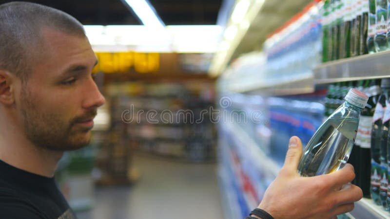Botella que elige y de compra del hombre joven de agua mineral en el supermercado Individuo que toma el producto de estantes en e