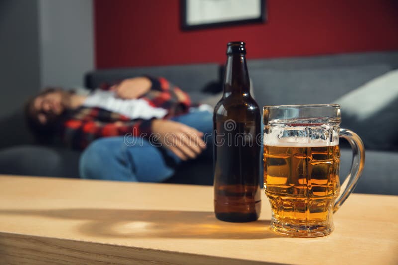 Botella y taza de cerveza en la tabla y el hombre borracho borroso que duermen en el sofá en casa Concepto del alcoholismo foto de archivo Botella y taza de cerveza en la tabla y el hombre borracho borroso que duermen en el sofá en casa Concepto del alcoholismo foto de archivo