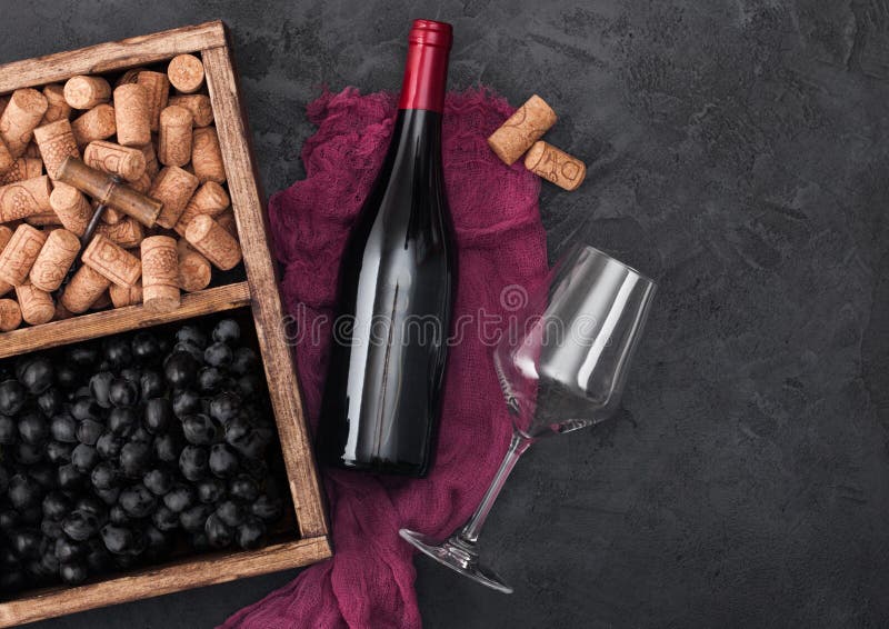 Bottle of red wine on red cloth with empty glass and dark grapes with corks and corkscrew inside vintage wooden box on dark wooden