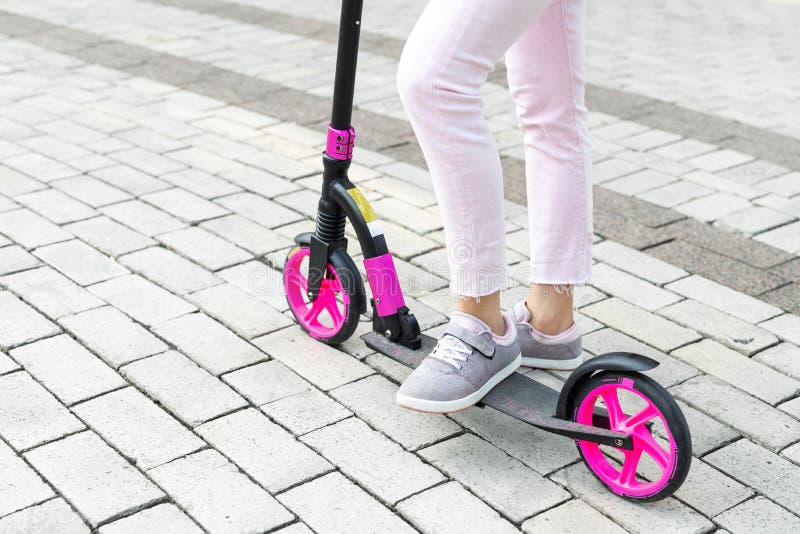Bottom part of young adult female person wearing pink casual jeans and sneakers riding push kick scooter at city street on summer stock photo Bottom part of young adult female person wearing pink casual jeans and sneakers riding push kick scooter at city street on summer stock photo