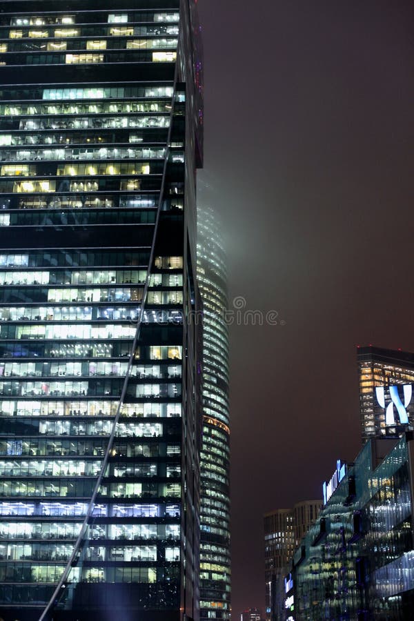 A bottom-up view of the facade of a modern office building at night in fog. Light of the night city. Moscow City