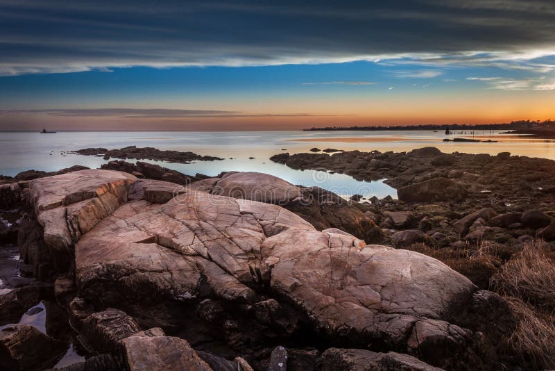Large rocks on the shore under a golden and blue sunset with a lighthouse in the background. Large rocks on the shore under a golden and blue sunset with a lighthouse in the background.