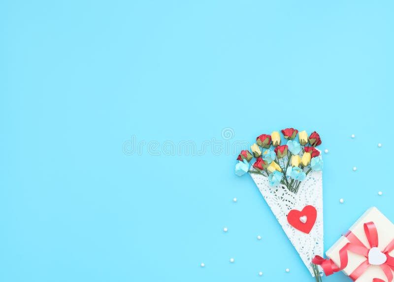 The bouquet of craft flowers wrapped in a white lace bundle and gift box with red ribbon on blue background with beads. Mother day
