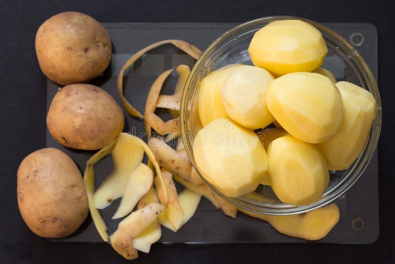 Bovenaanzicht van ongeschilde, rauwe aardappelen met schillen en gepelde aardappelen in glazen kom op glassnijplaat op zwarte ach royalty-vrije stock afbeeldingen Bovenaanzicht van ongeschilde, rauwe aardappelen met schillen en gepelde aardappelen in glazen kom op glassnijplaat op zwarte ach royalty-vrije stock afbeeldingen
