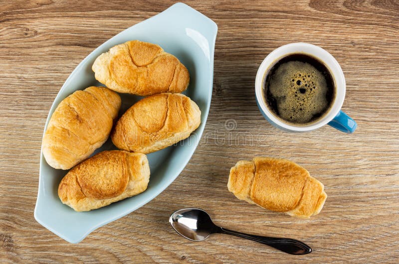 Bowl with small croissants, croissant, cup with black coffee, spoon on table. Top view