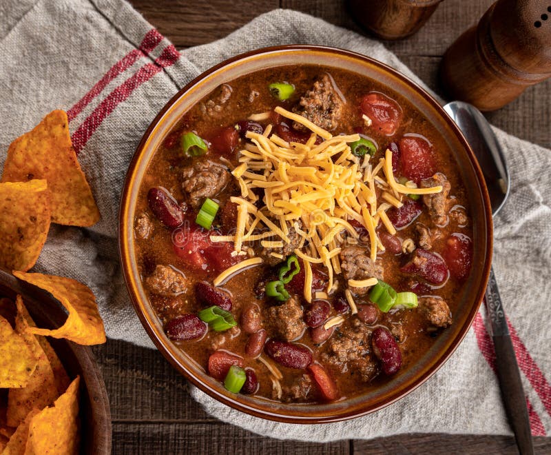 Bowl of chili con carne with shredded ceddar cheese and nacho chips on a rustic wooden table.  Overhead view. Bowl of chili con carne with shredded ceddar cheese and nacho chips on a rustic wooden table.  Overhead view