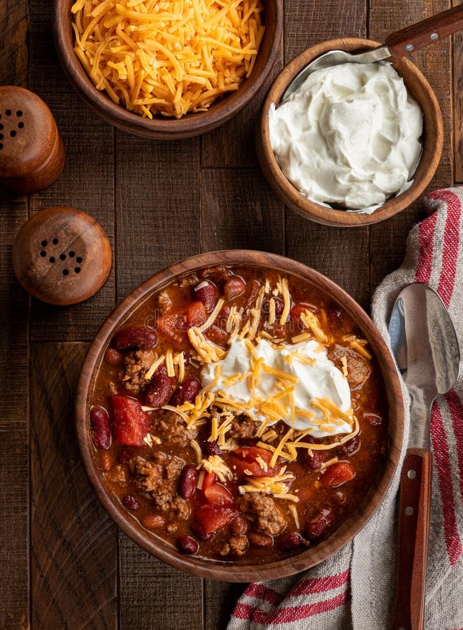 Bowl of chili con carne with shredded cheddar cheese and sour cream on a rustic wooden table.  Overhead view. Bowl of chili con carne with shredded cheddar cheese and sour cream on a rustic wooden table.  Overhead view