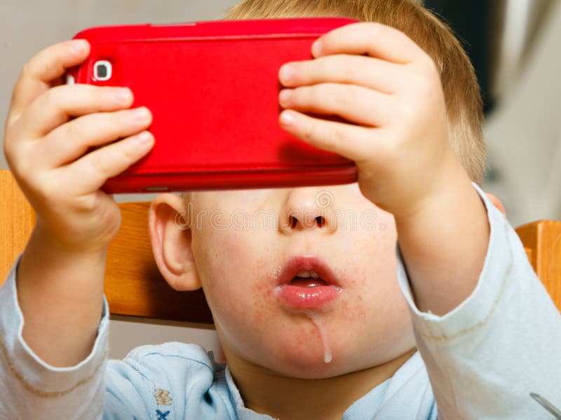 Technology generation. Happy childhood. Little boy child dirty mouth drooling, eating breakfast playing with mobile phone at table. Home. Technology generation. Happy childhood. Little boy child dirty mouth drooling, eating breakfast playing with mobile phone at table. Home.