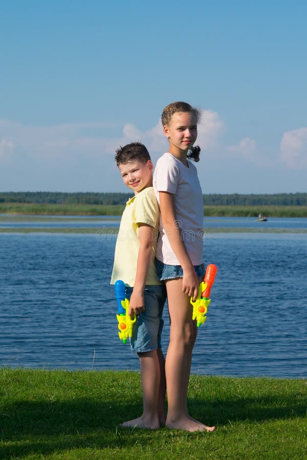 Boy and girl on the lake, holding water pistols and stand with their backs to each other, against a beautiful landscape royalty free stock photos Boy and girl on the lake, holding water pistols and stand with their backs to each other, against a beautiful landscape royalty free stock photos