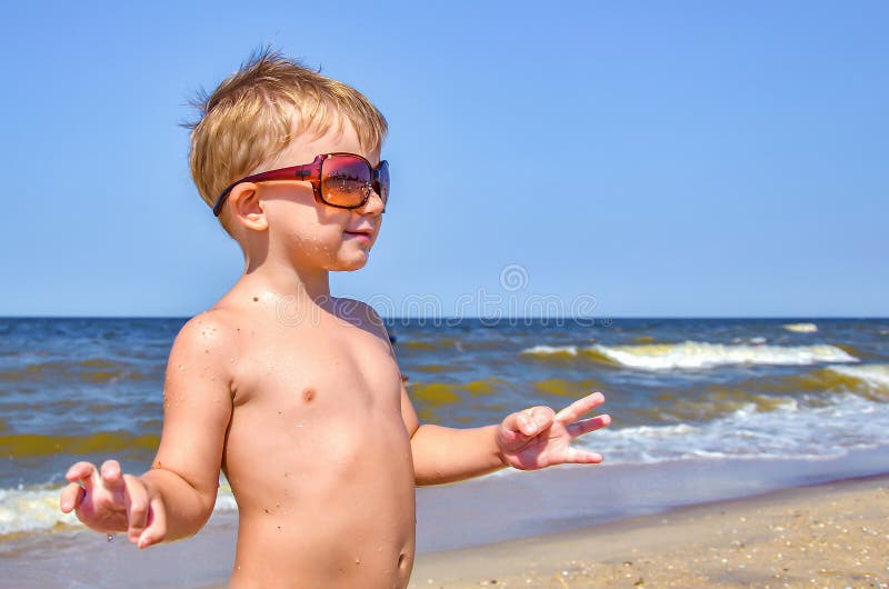 A boy in glasses stands on the beach by the sea