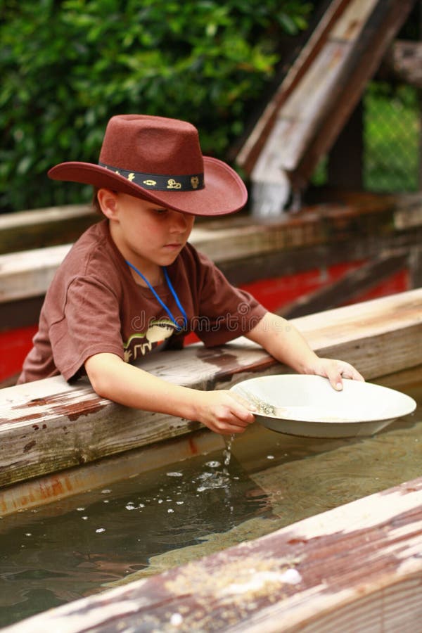 A seven year old boy pans for gold in a wooden trough. He wears a cowboy hat and holds a gold pan as he puts it in water to find gold nuggets. A seven year old boy pans for gold in a wooden trough. He wears a cowboy hat and holds a gold pan as he puts it in water to find gold nuggets.