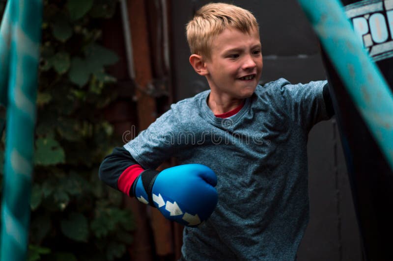 A Boy Punches a Punching Bag in Blue Gloves in the Yard Stock Photo