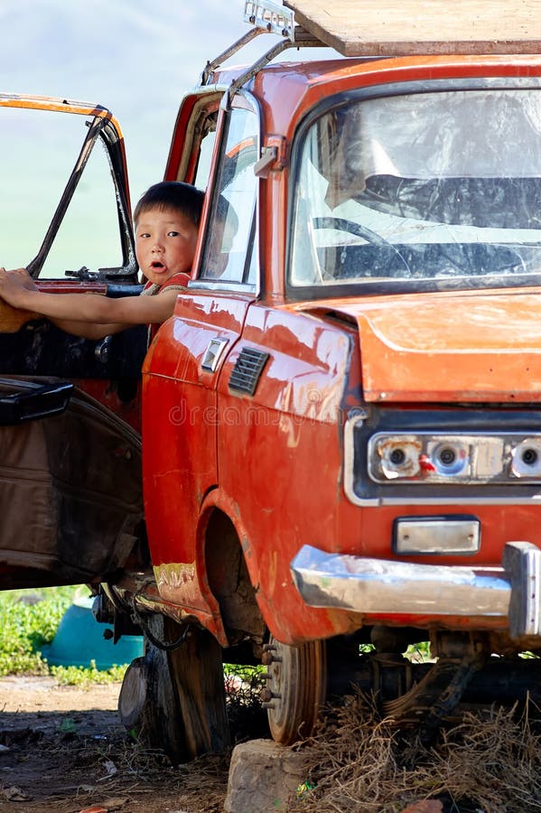 A boy is seated in the front seat of a wrecked car in Mongolia. He looks surprised while exploring the vehicle.