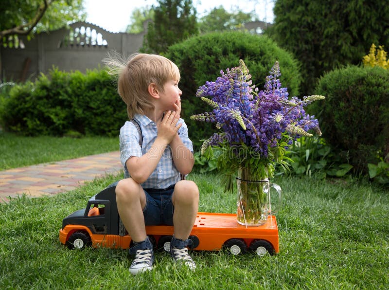 Boy in shorts and shirt sits on toy orange truck with bouquet of lupine flowers