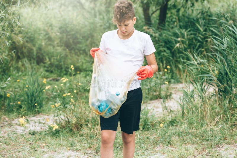 Boy in white t shirt in gloves collects garbage and plastic bottles into blue package on the beach. Young volunteer. Environment