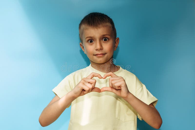 A boy in a yellow t-shirt on a blue background close-up with wide open eyes shows a heart on his fingers