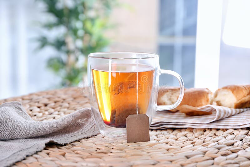 Brassage de la boisson chaude avec le sachet à thé dans la tasse en verre sur le tapis en osier image stock Brassage de la boisson chaude avec le sachet à thé dans la tasse en verre sur le tapis en osier image stock