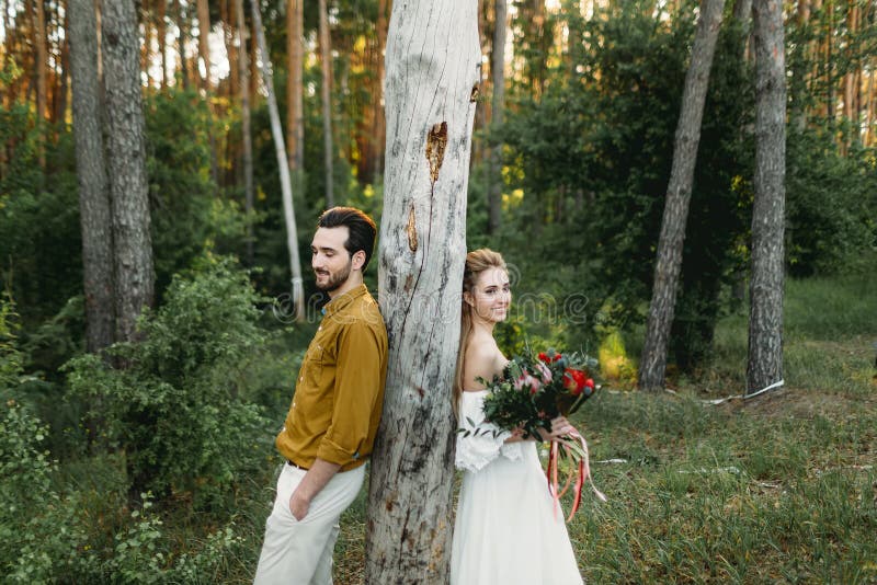 Bride and groom lean on the tree from different sides. Newlyweds are walking in the forest. Artwork,. Bride and groom lean on the tree from different sides. Newlyweds are walking in the forest. Artwork,