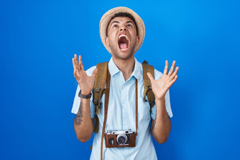 Brazilian young man holding vintage camera crazy and mad shouting and yelling with aggressive expression and arms raised. frustration concept. Brazilian young man holding vintage camera crazy and mad shouting and yelling with aggressive expression and arms raised. frustration concept