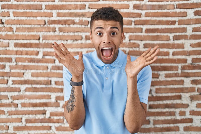 Brazilian young man standing over brick wall celebrating crazy and amazed for success with arms raised and open eyes screaming excited. winner concept. Brazilian young man standing over brick wall celebrating crazy and amazed for success with arms raised and open eyes screaming excited. winner concept