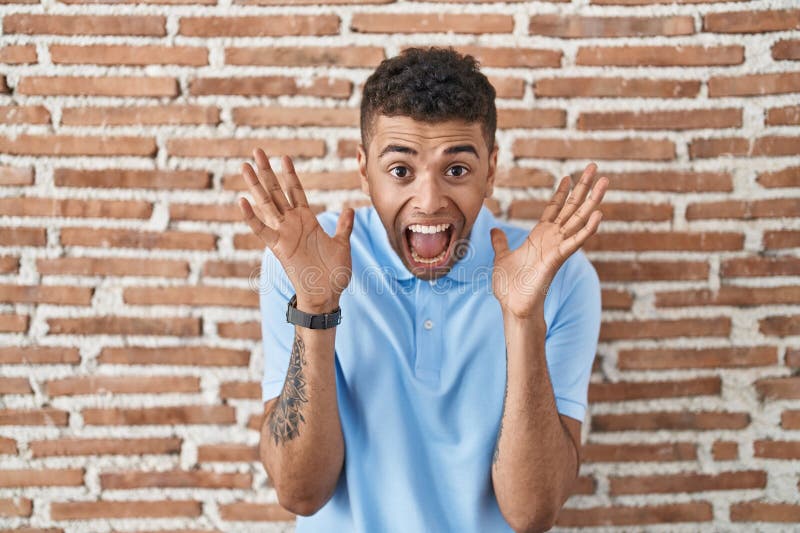 Brazilian young man standing over brick wall celebrating mad and crazy for success with arms raised and closed eyes screaming excited. winner concept. Brazilian young man standing over brick wall celebrating mad and crazy for success with arms raised and closed eyes screaming excited. winner concept