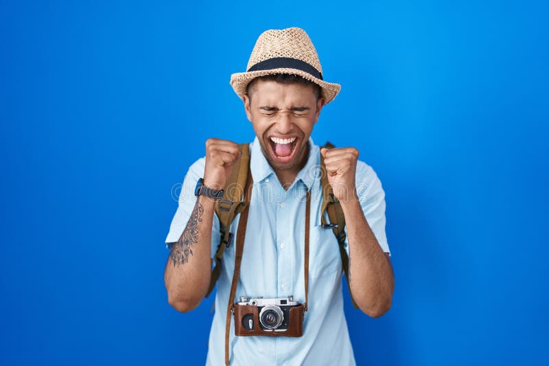 Brazilian young man holding vintage camera excited for success with arms raised and eyes closed celebrating victory smiling. winner concept. Brazilian young man holding vintage camera excited for success with arms raised and eyes closed celebrating victory smiling. winner concept