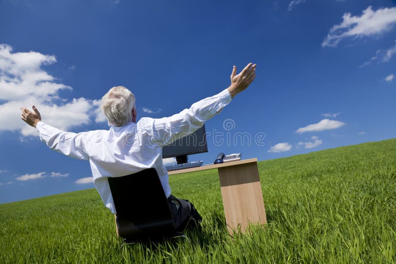 Business concept shot showing an older male executive arms raised using a computer in a green field with a blue sky complete with fluffy white clouds. Shot on location not in a studio. Business concept shot showing an older male executive arms raised using a computer in a green field with a blue sky complete with fluffy white clouds. Shot on location not in a studio.