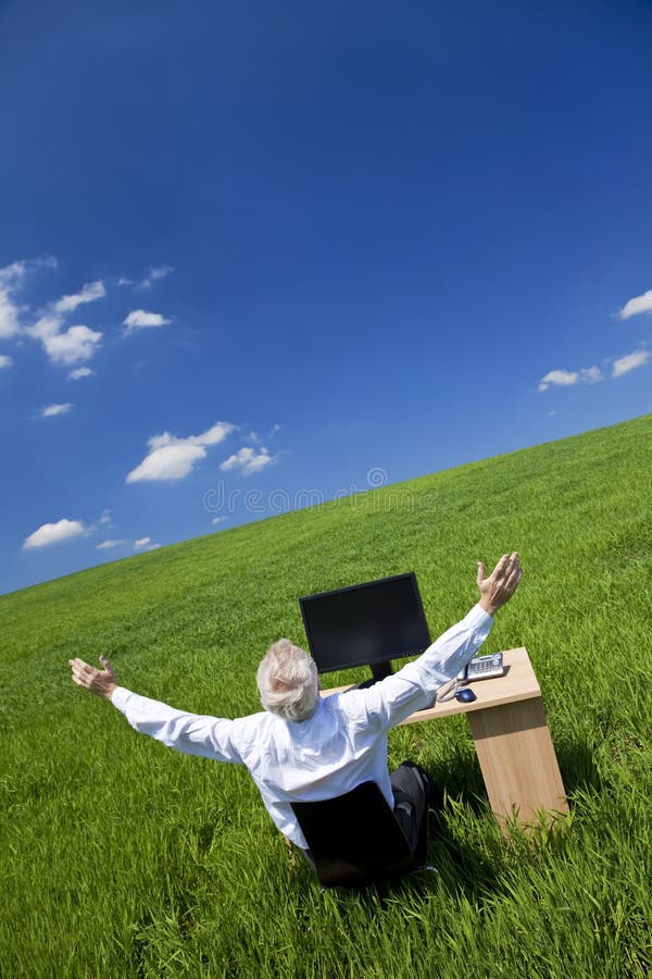 Business concept shot showing an older male executive arms raised using a computer in a green field with a blue sky complete with fluffy white clouds. Shot on location not in a studio. Business concept shot showing an older male executive arms raised using a computer in a green field with a blue sky complete with fluffy white clouds. Shot on location not in a studio.