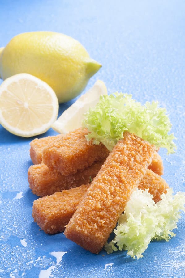 Breaded fritou dedos de peixes com o tomate do vermelho da alface e de cereja com fatia do limão no fundo azul com respingo da ág fotografia de stock royalty free Breaded fritou dedos de peixes com o tomate do vermelho da alface e de cereja com fatia do limão no fundo azul com respingo da ág fotografia de stock royalty free