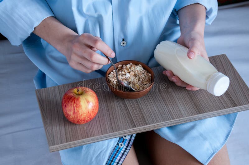 Breakfast in bed, a girl in a blue shirt sitting on a white sheet and Breakfast cereal with milk. Girl holding a bottle of milk. on a tray a bowl of porridge and a red Apple.