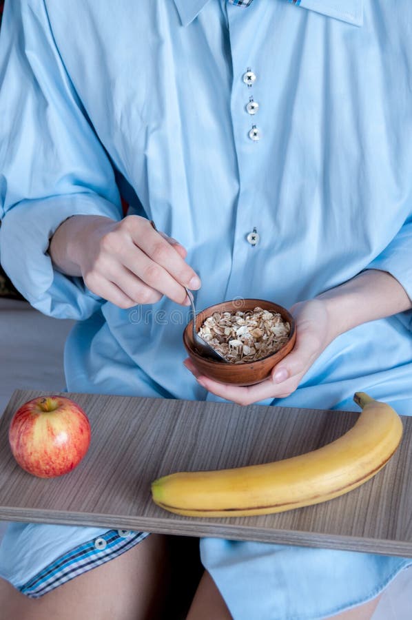 Breakfast in bed, a girl in a blue shirt sitting on a white sheet and Breakfast cereal with milk. Girl holding a Cup of porridge. On a tray banana and red Apple, healthy diet