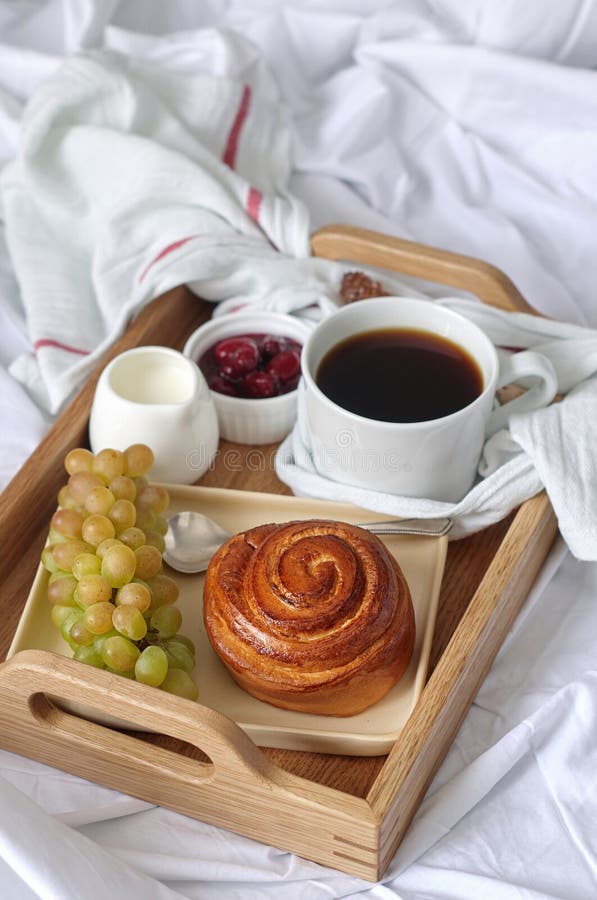 Breakfast on white bed sheets in hotel room. Close up of wooden tray with coffee, bun, jam and grape.