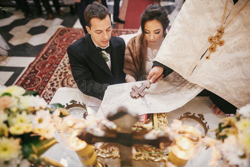 Bride and groom pronouncing vows, holding hands on bible on altar and priest holding golden cross on hands during holy matrimony in church