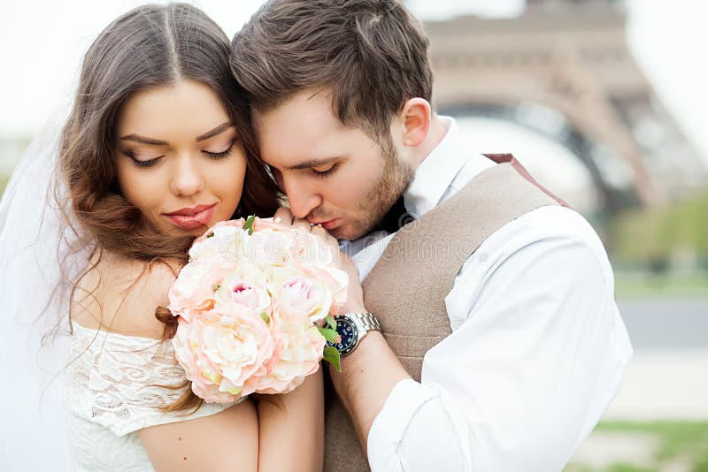 Bride and groom sitting on bench in park, holding hands of each other and bouquet. Groom holding his head on bride`s shoulder and