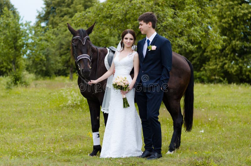 Bride and groom are standing in the park near the horse, wedding walk. White dress, happy couple with an animal. Green background