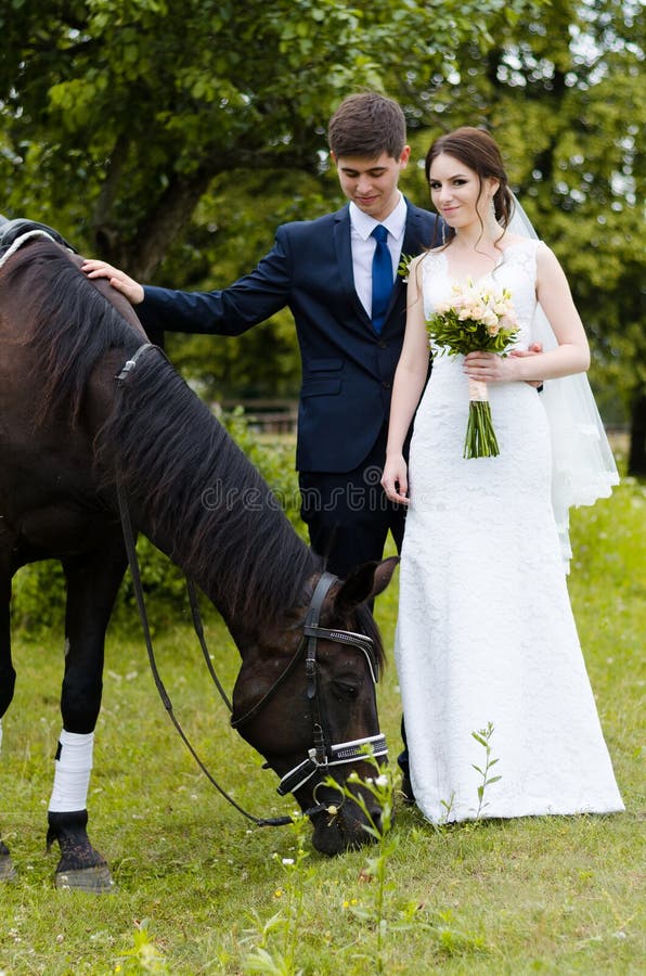 Bride and groom are standing in the park near the horse, wedding walk. White dress, happy couple with an animal. Green background