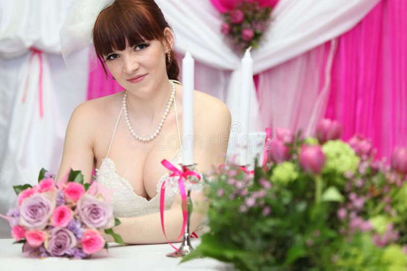 Happy bride wearing white dress sits at table with candles and holds bouquet of roses