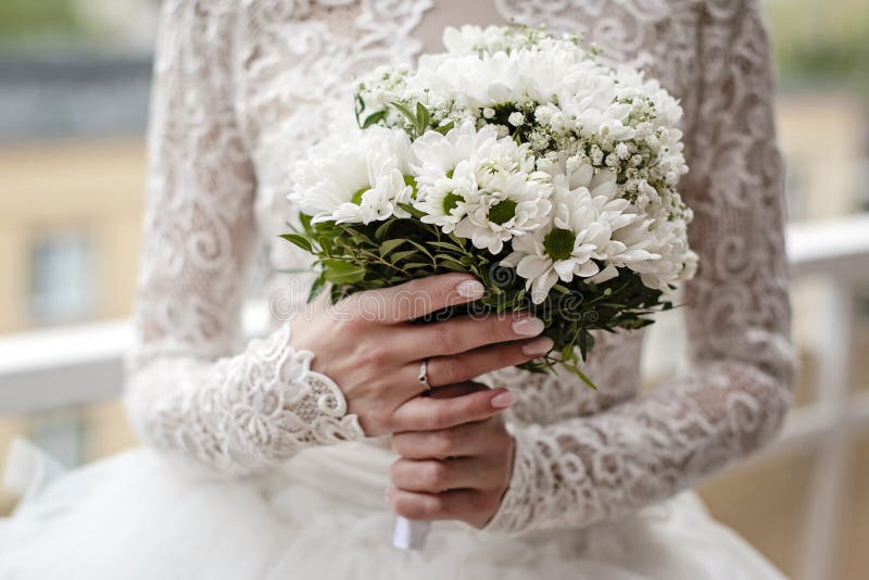 Bride in white lace dress is holding wedding flowers. Close-up on brideâ€™s hands wearing engagement ring and holding white