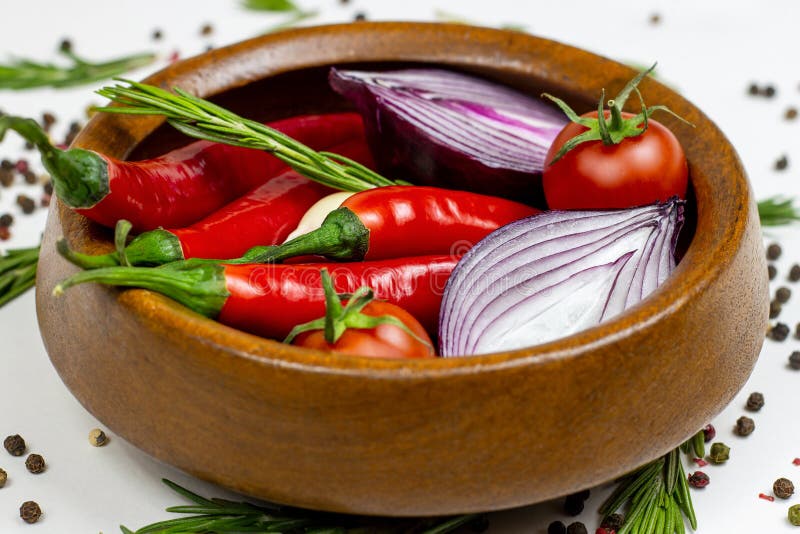 Bright summer vegetables: onion, garlic, tomato, red chili pepper, rosemary in brown wooden bowl and peppercorns on white table