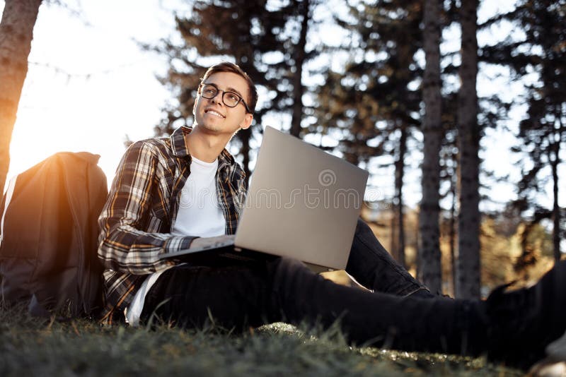 Bright young male student in casual outfit using notebook or laptop while sitting on the grass at the park