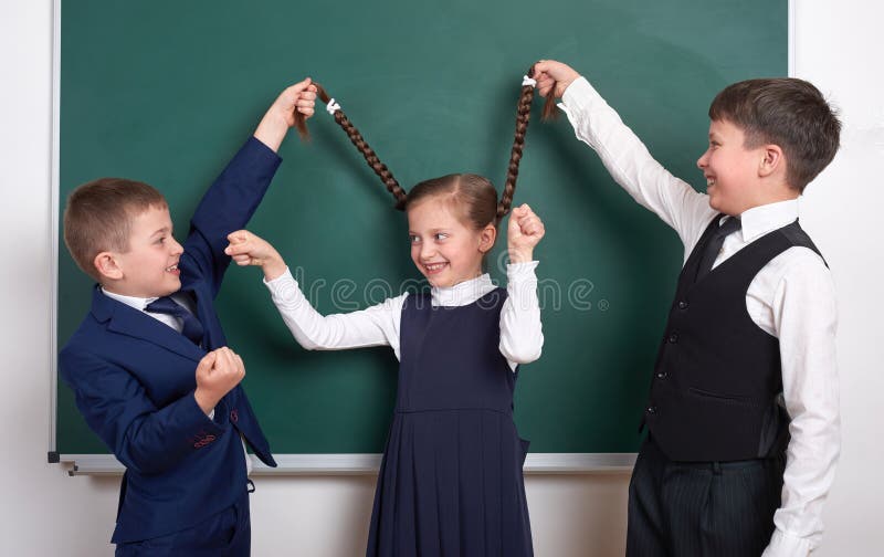 A brincadeira e o divertimento ter, meninos puxam as tranças da menina, perto do fundo vazio do quadro da escola, vestido no tern fotos de stock A brincadeira e o divertimento ter, meninos puxam as tranças da menina, perto do fundo vazio do quadro da escola, vestido no tern fotos de stock