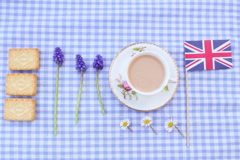 A cup of milky tea with traditional custard cream type biscuits (not branded) with a union jack flag on a gingham tablecloth. A cup of milky tea with traditional custard cream type biscuits (not branded) with a union jack flag on a gingham tablecloth